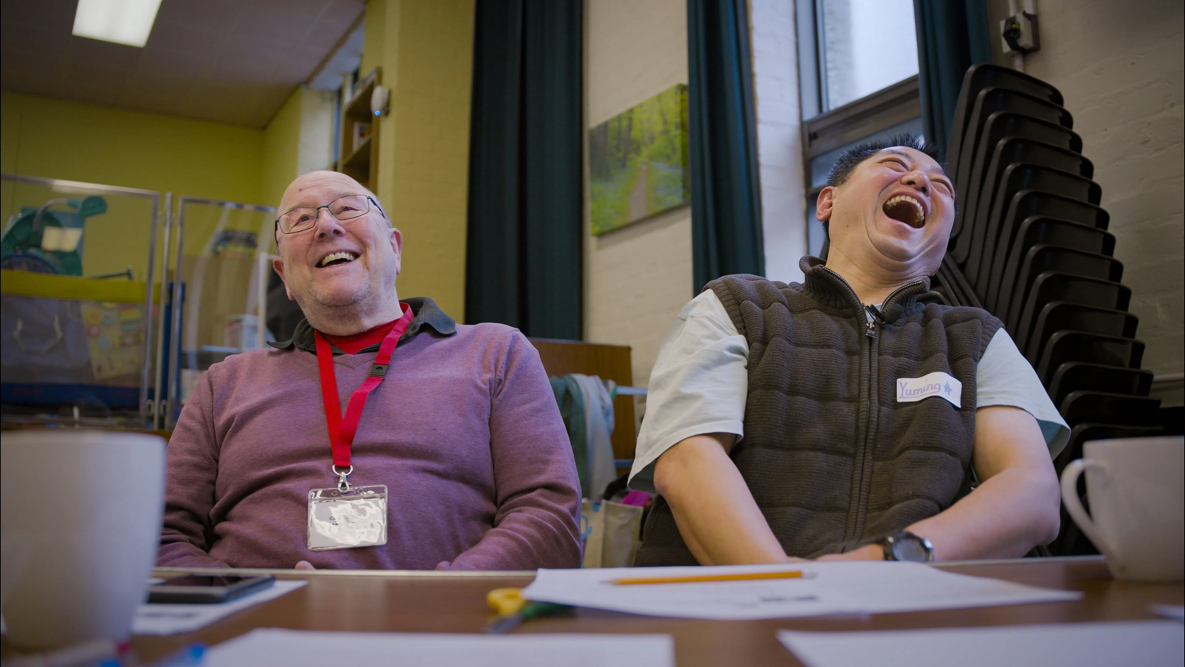 Two people sit at a table, laughing heartily. Papers and mugs are on the table, and chairs are stacked in the background, creating a joyful atmosphere.